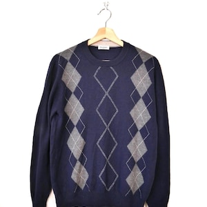 May include: A navy blue sweater with a grey argyle pattern. The sweater is suspended on a wooden hanger against a white backdrop. The argyle design features diamond shapes in a lighter grey colour.