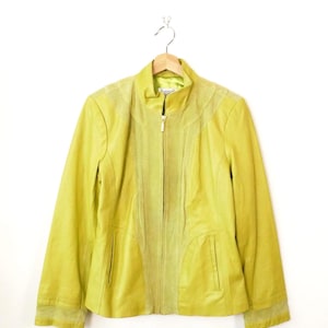 Peut inclure: Veste en cuir chartreuse avec fermeture éclair et col montant. La veste présente des détails perforés sur les épaules et un panneau en daim au centre. La veste est suspendue à un cintre en bois.
