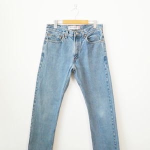 May include: Light blue denim jeans hanging on a wooden hanger. The jeans feature a classic five-pocket design, a button and zip closure, and a straight leg fit. The jeans are a medium wash and appear pre-worn.