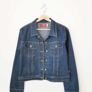 May include: A dark blue denim jacket with a button-up front, two chest pockets, and a small red logo on the sleeve. The jacket is suspended on a wooden hanger against a white backdrop.