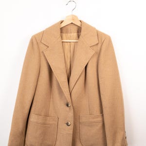 Könnte beinhalten: Ein beigefarbener Blazer mit steigendem Revers, zwei aufgesetzten Vordertaschen und einem Zwei-Knopf-Verschluss. Das Kleidungsstück ist mit langen Ärmeln und einem sichtbaren Innenfutter ausgestattet und hängt auf einem Holzkleiderbügel.