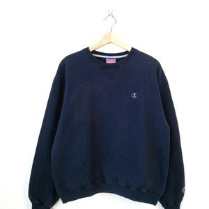May include: A navy blue long-sleeve crewneck sweatshirt featuring a small embroidered Champion logo on the left chest and a larger embroidered Champion logo on the left sleeve cuff. The garment has a ribbed collar, cuffs, and hem.