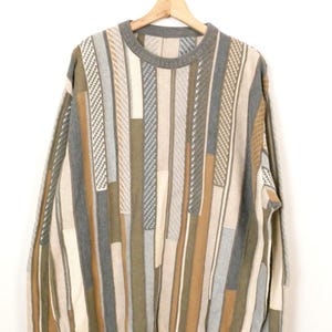 Puede incluir: Un su&eacute;ter vintage con cuello redondo y mangas largas. El su&eacute;ter presenta un patr&oacute;n de rayas verticales en tonos grises, beige y marr&oacute;n. Cuelga de una percha de madera sobre un fondo blanco.