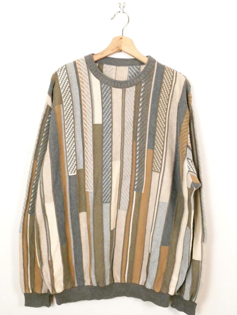 Puede incluir: Un su&eacute;ter vintage con cuello redondo y mangas largas. El su&eacute;ter presenta un patr&oacute;n de rayas verticales en tonos grises, beige y marr&oacute;n. Cuelga de una percha de madera sobre un fondo blanco.