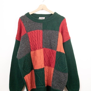 Könnte beinhalten: Ein langärmeliger Pullover mit Rundhalsausschnitt und einem Patchwork-Design aus dunkelgrünen, anthrazitfarbenen, roten und orangefarbenen Strickbahnen. Das Kleidungsstück zeigt verschiedene Texturen wie Zopfmuster, Rippstrick und glatte Muster.