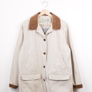 May include: A beige L.L. Bean jacket with a brown corduroy collar and cuffs. The jacket has a button-up front, two chest pockets, and two lower pockets. The jacket is hanging on a wooden hanger.