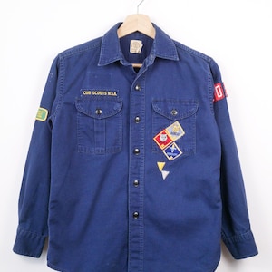 Könnte beinhalten: Ein blaues Vintage-Hemd der Cub Scouts B.S.A. mit langen Ärmeln und Knopfleiste. Das Hemd hat zwei Brusttaschen und mehrere Aufnäher, darunter ein Bobcat-Aufnäher und ein rot-weißer Aufnäher am rechten Ärmel. Der Text "CUB SCOUTS B.S.A." ist auf der linken Brusttasche gestickt.