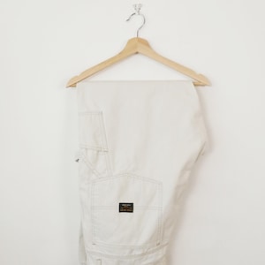 May include: Off-white work pants hanging on a wooden hanger. The pants have multiple pockets, visible stitching, and a small rectangular label on the back pocket. The pants are made of a durable material.