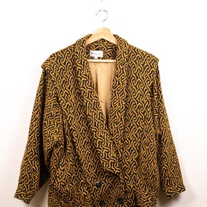 May include: A double-breasted blazer featuring a mustard yellow and black interlocking geometric pattern. The jacket has a wide shawl collar, long sleeves with cuffs, and two black buttons at the front. A label with the text "Sanda" is visible.