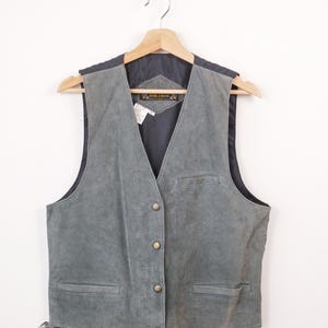 May include: A grey suede waistcoat featuring a V-neckline, four metallic buttons down the front, and two lower welt pockets. The interior is lined with dark grey fabric and includes a label with the text Don Taylor. It hangs on a wooden hanger.