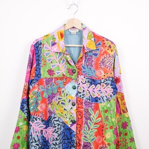 Peut inclure: Veste patchwork colorée avec un col et des manches longues. La veste présente divers motifs floraux et cachemire dans les tons de rose, vert, bleu et orange. Elle est boutonnée et suspendue à un cintre en bois.