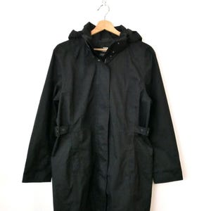 May include: Black hooded jacket with a button-up front and belt loops. The jacket is hanging on a wooden hanger. The jacket has a hood and long sleeves.