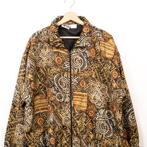 May include: A zip-up jacket featuring a complex, multi-coloured pattern in shades of brown, black, tan, and white. The garment has a high collar, long sleeves with elastic cuffs, and a black zip closure. A Kaktus brand label is visible at the neck.
