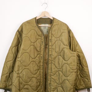 Peut inclure: Une veste doublure matelassée vert olive avec un motif de surpiqûres ondulées, une encolure sans col et une fermeture boutonnée à l'avant. L'intérieur présente une étiquette rectangulaire avec du texte imprimé et les manches ont des pattes.