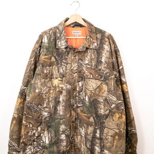Peut inclure: Une chemise à manches longues Carhartt boutonnée avec un motif camouflage forestier dans les tons marron, beige et vert. La chemise présente une doublure intérieure matelassée orange vif, un col classique, deux poches poitrine à rabat et des boutons en métal.