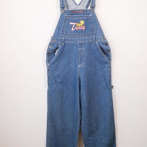 May include: Blue denim overalls featuring a front bib pocket with an embroidered Tweety Bird graphic and the text Tweety in pink script. The garment includes adjustable shoulder straps with metal buckles, side button closures, and wide legs.