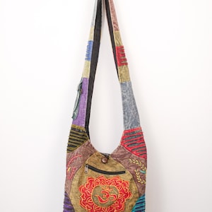 May include: A patchwork crossbody bag featuring a variety of coloured fabric panels including brown, purple, blue, and red. The front displays a prominent red embroidered Om symbol, a zippered pocket, and decorative stitching throughout the exterior.