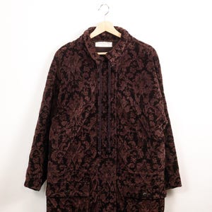 Peut inclure: Une veste marron foncé et noire avec un motif floral damassé répétitif. La veste présente un col montant, des manches longues, deux grandes poches avant à rabat et un cordon de serrage vertical central de style corde au niveau du col.