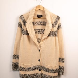 May include: A cream-colored knit cardigan featuring a shawl collar, long sleeves, and four dark buttons down the front. The garment is decorated with horizontal black and white speckled stripes across the chest, sleeves, and hem, and includes two front pockets.