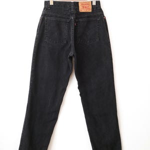 Op de afbeelding: Zwarte spijkerbroek van denim met een hoge taille, twee achterzakken met kenmerkende gebogen stiksels, een klein rood label op de rechterzak en een beige merklabel van imitatieleer op de tailleband. De broek hangt aan een metalen kledinghanger.