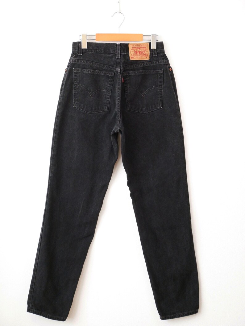 Op de afbeelding: Zwarte spijkerbroek van denim met een hoge taille, twee achterzakken met kenmerkende gebogen stiksels, een klein rood label op de rechterzak en een beige merklabel van imitatieleer op de tailleband. De broek hangt aan een metalen kledinghanger.