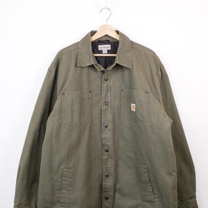 May include: Olive green work jacket with a collared neckline and multiple pockets. The jacket has a small Carhartt logo patch on the chest pocket. It is hanging on a wooden hanger against a white background.