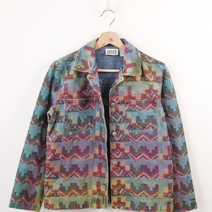 May include: A long-sleeve denim jacket featuring a multi-coloured geometric pattern in shades of teal, purple, red, and yellow. The jacket includes a pointed collar, a button-front closure, and two chest pockets. A Chico's Design label is visible.