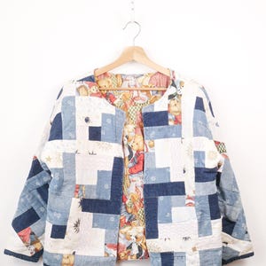May include: A reversible jacket featuring a patchwork exterior of blue denim and white fabric squares with snowflake patterns. The interior lining displays a colourful print of teddy bears and various small objects. The jacket has a collarless neckline.