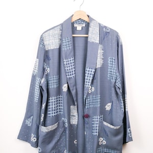 May include: A slate blue blazer featuring a patchwork-style print with white grid patterns, floral motifs, and small dots. This long-sleeve jacket has a shawl collar, two front patch pockets, and a single dark purple button closure at the centre front.