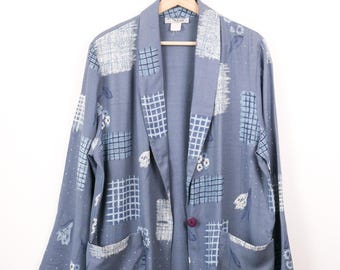 Pode incluir: Um blazer azul ardósia com uma estampa estilo patchwork, apresentando padrões de grade brancos, motivos florais e pequenos pontos. Esta jaqueta de manga comprida possui gola xale, dois bolsos frontais e um botão roxo escuro central.