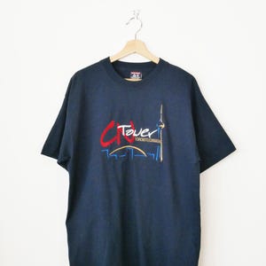 May include: Dark blue t-shirt featuring a graphic of the CN Tower and the word "Tower" in red and blue. The text "Toronto Canada" is also visible. The shirt is displayed on a wooden hanger.