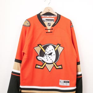 May include: An orange Reebok Anaheim Ducks hockey jersey featuring a large duck mask logo on the chest, crossed hockey sticks, black and gold sleeve stripes, a black collar with laces, and a size S tag on the lower front hem.