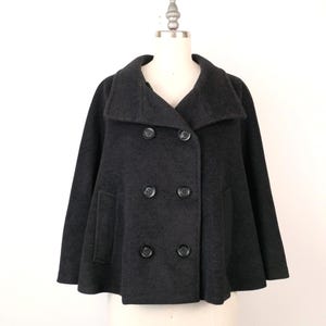 Peut inclure: Un manteau cape gris anthracite avec un devant croisé et un grand col. Le manteau comporte quatre boutons foncés et deux poches. Le vêtement est présenté sur un mannequin contre un fond blanc.