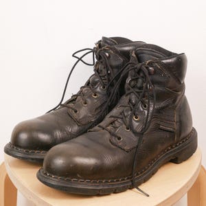 May include: A pair of black leather work boots featuring a rounded toe, a lace-up closure with metal eyelets, a padded collar, and a thick, durable rubber sole. The boots have visible stitching along the welt and a textured, worn-in leather finish.
