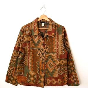 May include: A patterned button-down jacket in shades of brown, orange, and green, hanging on a wooden hanger. The geometric design features diamond and zigzag shapes. The jacket is displayed against a white backdrop.