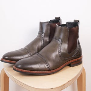 Peut inclure: Paire de bottines Chelsea en cuir vert olive avec empiècements latéraux élastiques marron et perforations décoratives. Les bottines ont un bout arrondi et une semelle marron avec un bord marron clair. Elles sont posées sur un tabouret en bois clair.