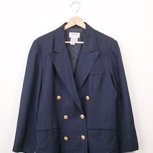 May include: A navy blue double-breasted blazer featuring six gold-toned buttons, notched lapels, and two front welt pockets. The garment is displayed on a wooden hanger and includes a white interior label with the text Talbots Petite.