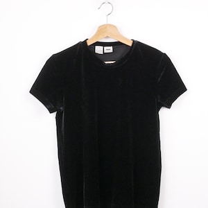 May include: A black velvet t-shirt with short sleeves hangs from a wooden hanger. The shirt has a round neckline and a soft, textured appearance. The tag reads "DKNY".