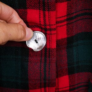 May include: Close-up of a red, black, and green plaid fabric with a silver button. The button is being held in place. The fabric has a classic lumberjack or flannel pattern.