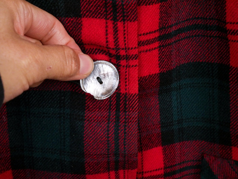 May include: Close-up of a red, black, and green plaid fabric with a silver button. The button is being held in place. The fabric has a classic lumberjack or flannel pattern.