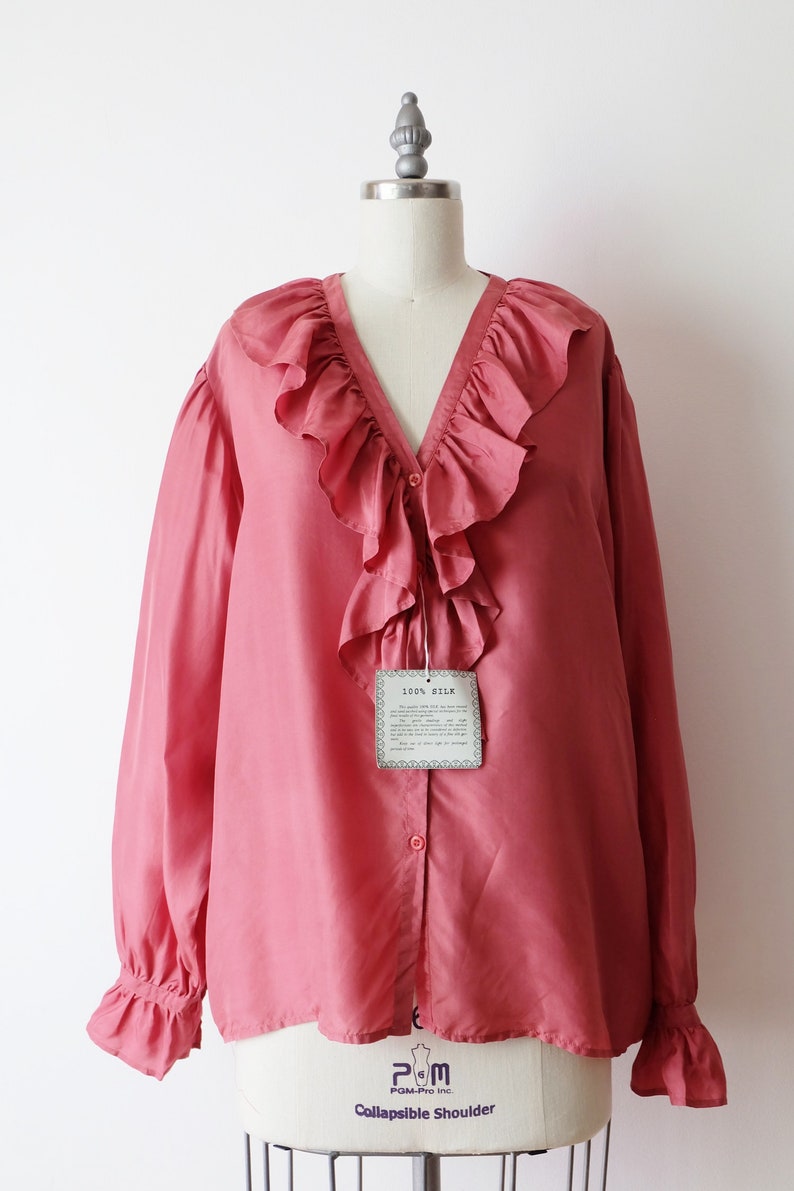 May include: A salmon pink silk blouse with a large ruffled collar and long sleeves. The blouse has a button-up front and a tag that reads "100% Silk".