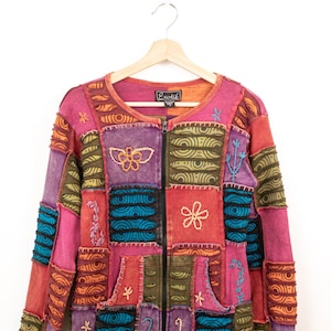 Può includere: Una giacca a maniche lunghe con cerniera e design patchwork nei toni del rosa, viola, arancione e verde acqua. Il capo presenta motivi floreali e astratti ricamati, cuciture a vista e due tasche frontali. L'etichetta al collo riporta Exotik.