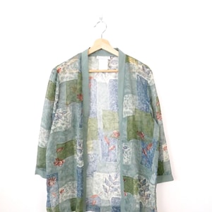 May include: A sheer, open-front kimono-style top in shades of blue, green, and white, with a patchwork design. The fabric features floral and leaf patterns. The top is hanging on a wooden hanger against a white background.