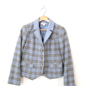 May include: A light blue and grey plaid blazer featuring a solid light blue notched collar, three front buttons, and two flap pockets. The interior label reads Pendleton. The garment is displayed on a wooden hanger against a plain white background.