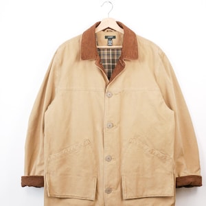 May include: A tan jacket with a brown corduroy collar and cuffs, hanging on a wooden hanger. The jacket has a button-up front, two large front pockets, and a plaid lining visible at the collar. The jacket is a neutral color.