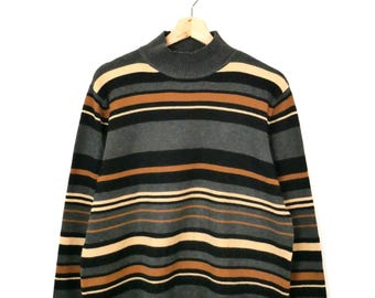 May include: A long-sleeve mock neck sweater featuring horizontal stripes in shades of charcoal gray, black, and tan. The garment is constructed with a ribbed collar and a straight hemline finished with a fringe of hanging yarn strands.