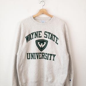 May include: A light gray crewneck sweatshirt featuring the dark green text "WAYNE STATE UNIVERSITY" arched over a shield logo with a white "W" inside. The garment has long sleeves, ribbed cuffs, and a small Champion logo on the left sleeve.