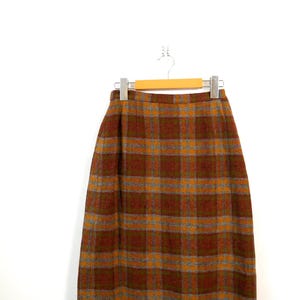 May include: A vintage knee-length skirt with a plaid pattern in shades of brown, orange, and grey. The skirt is hanging on a wooden hanger against a white background. The fabric appears to be a woven material.