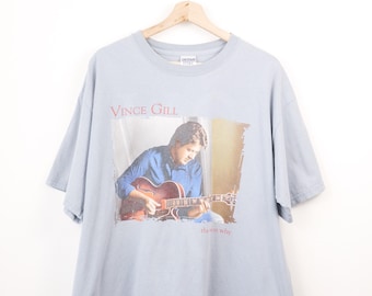 May include: A light blue short-sleeve t-shirt featuring a graphic print of Vince Gill playing a hollow-body electric guitar. The text "VINCE GILL" appears in red above the image, and the text "the son why" is printed in red below the image.