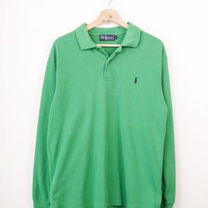 May include: A long-sleeve polo shirt in a vibrant green color featuring a dark blue embroidered logo on the left chest. The shirt has a classic collar, a two-button placket, and a label at the neck that reads Ralph Lauren. It is displayed on a hanger.